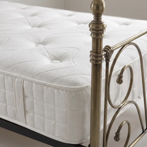 A close-up of a white tufted mattress on a bed frame with an antique brass metal headboard and footboard.