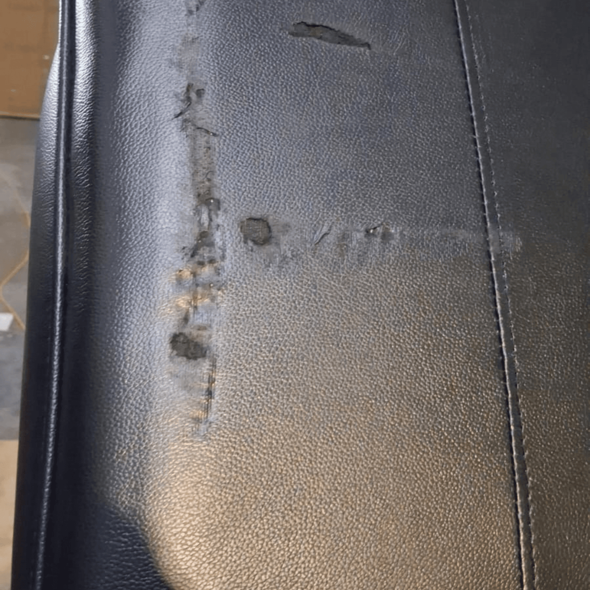 Close-up of a black textured leather or faux-leather surface with visible scratches, scuffs, and wear marks along a vertical seam.